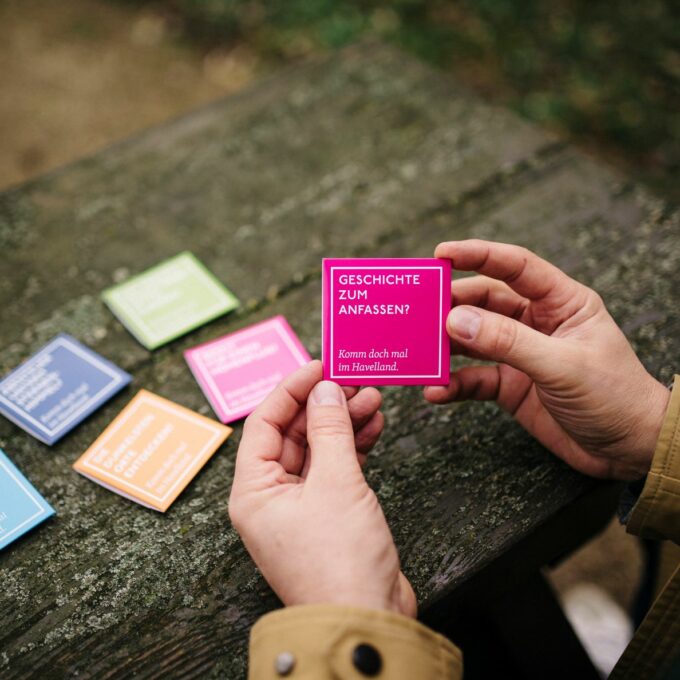 Männerhände halten pinke Kondomverpackung mit Aufdruck: Geschichte zum Anfassen? Komm doch mal im Havelland.