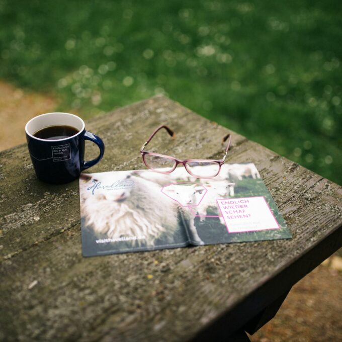 Brillenputztuch Havelland mit Schafsmotiv und Spruchaufdruck "Endlich wieder Schaf sehen? Komm doch mal ins Havelland." liegt auf dem Tisch neben rosa Brille und blauer Kaffeetasse